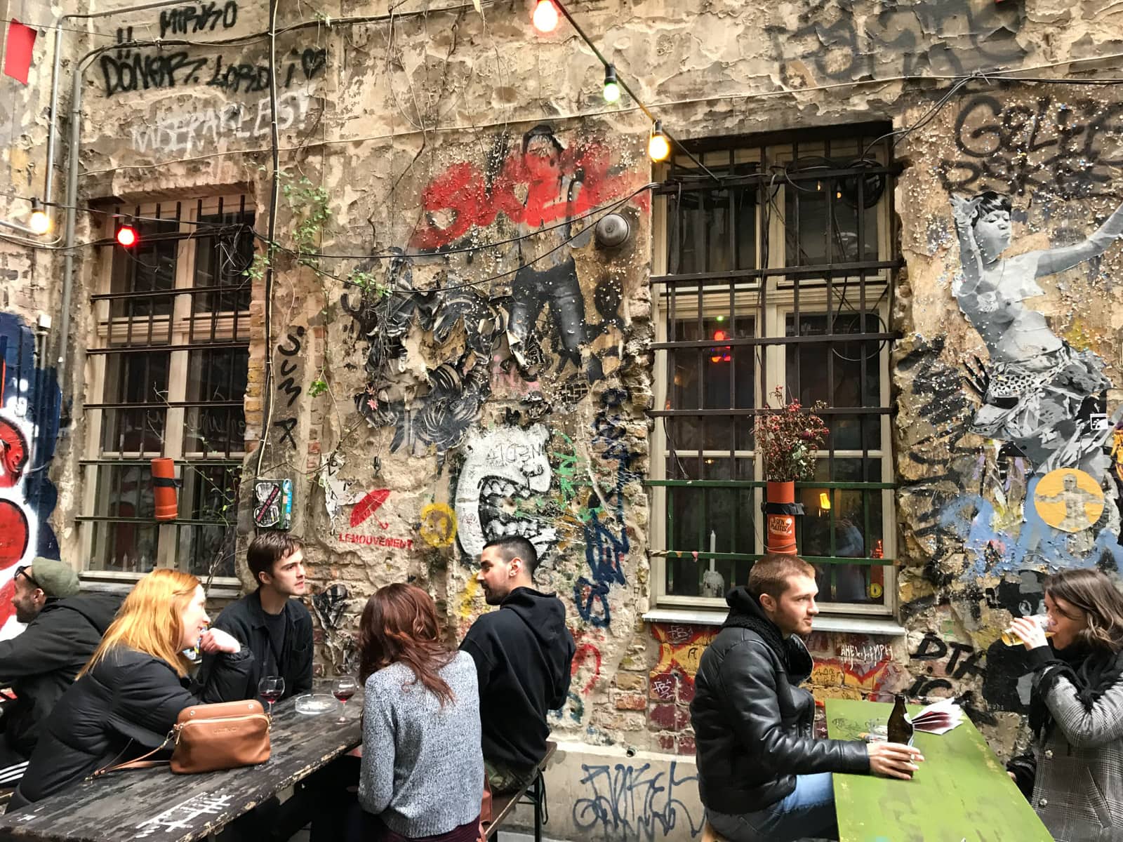 Some people sitting at tables in a courtyard, behind them a building covered in graffiti.