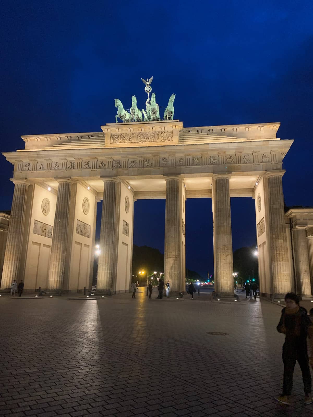 A very tall arch gate with many pillars, and a statue of four horses on top. It’s nighttime and the arch looks lit up.