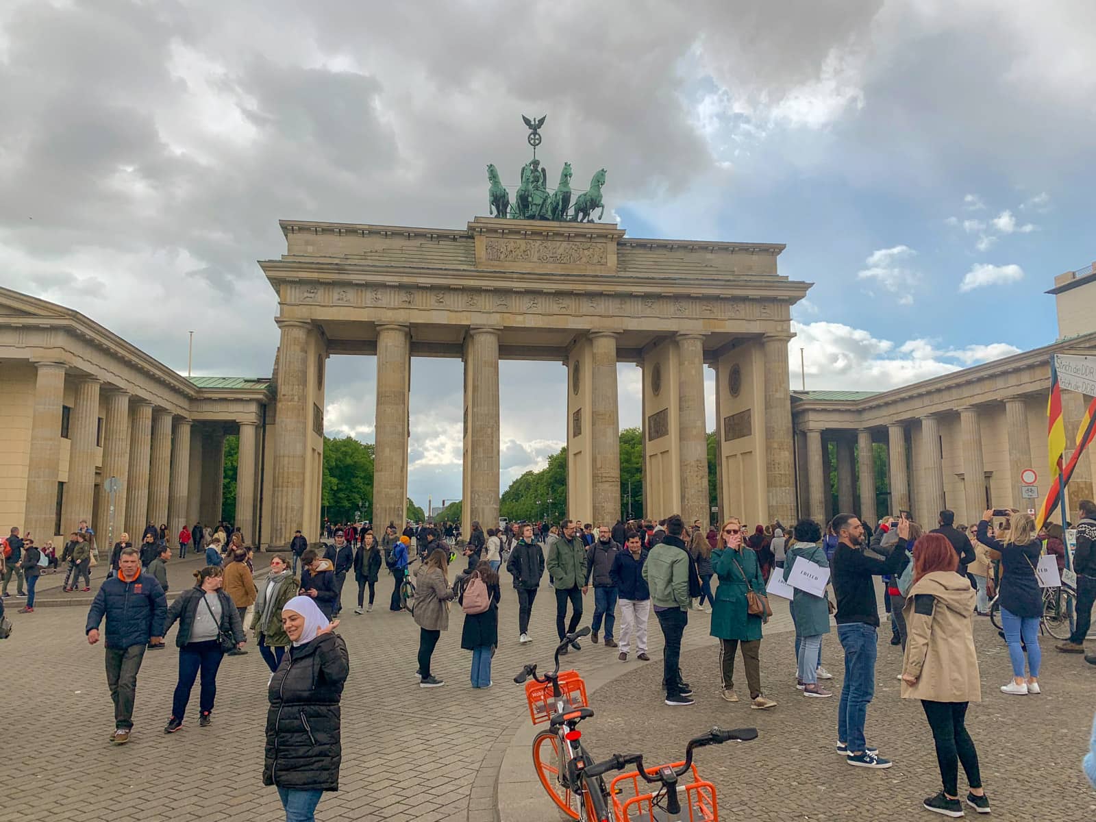 A big arch gate with a statue of four horses on top, during the daytime. There is a small crowd of people taking photos. The sky has many clouds.