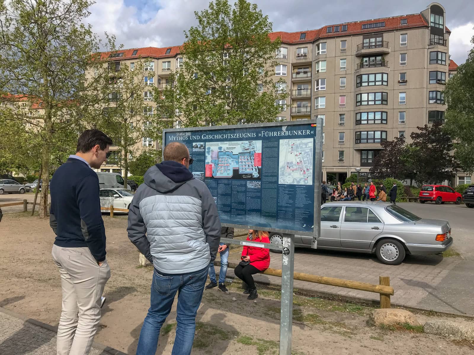 Two men standing by a sign in front of an open carpark. The men are reading the sign, which has detailed information about Hitler’s bunker