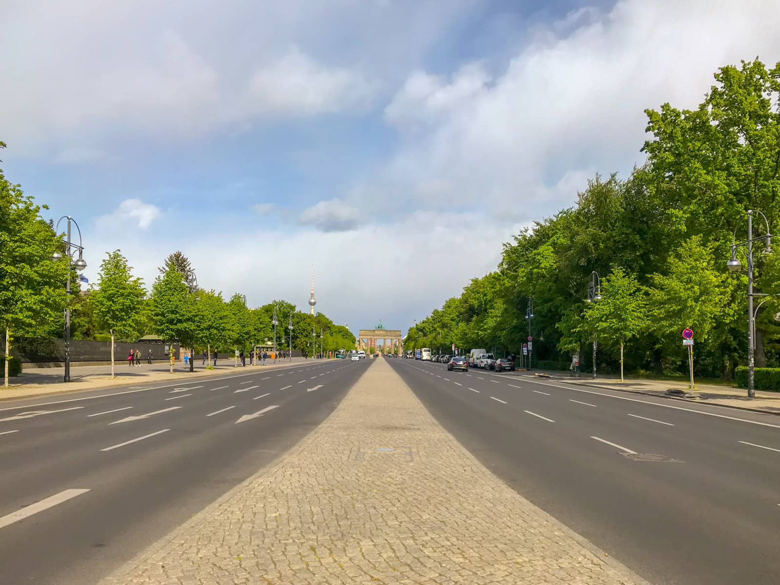 A view from the middle of a road, with road lanes on either side of the median strip, and bright green trees outside of the road. There is a tall arch pillared gate in the distance