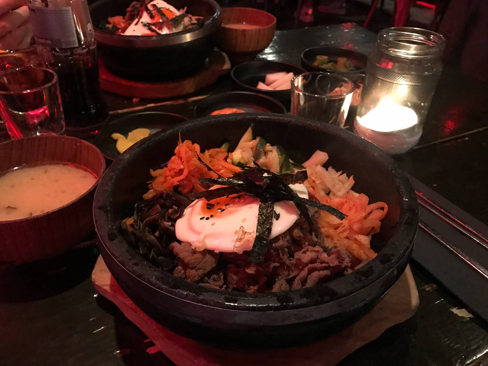 A hot stone pot on a wooden plate, in a restaurant. There is also some soup and drinks placed on the table