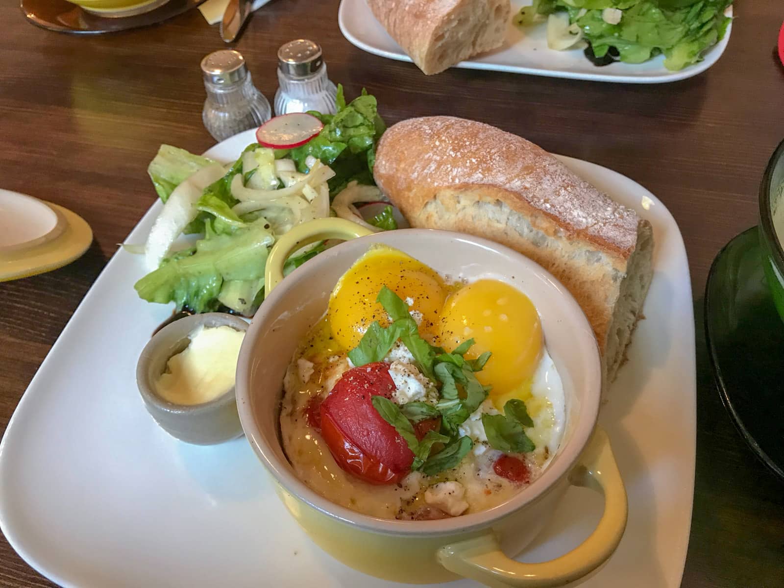A white plate with ciabatta bread, salad, butter in a small cup, and a small pot of baked eggs