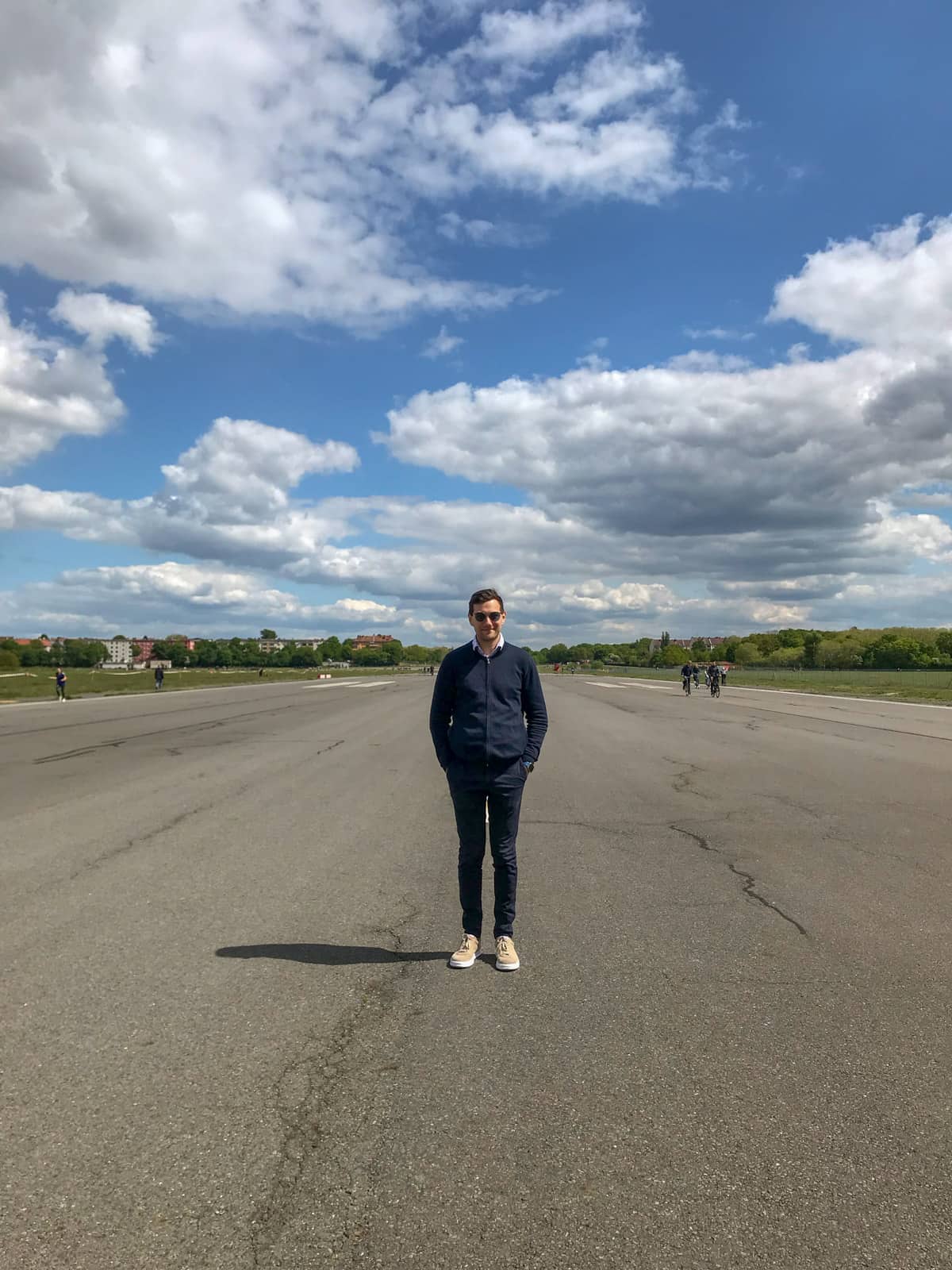 A man dressed in a navy jacket and pants, wearing sunglasses, with his hands in his pockets. He is standing in an open concrete area. The sky is very blue with many clouds.