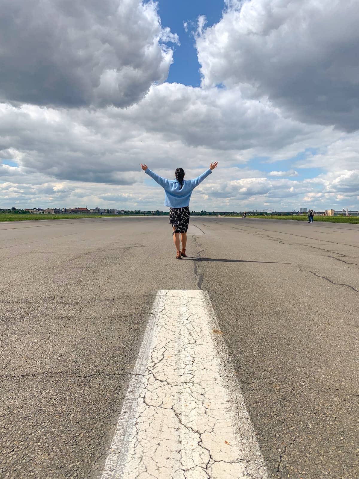 A woman seen from behind, with her arms in the air. She’s standing on an open stretch of concrete, and the sky is very blue with a lot of clouds.