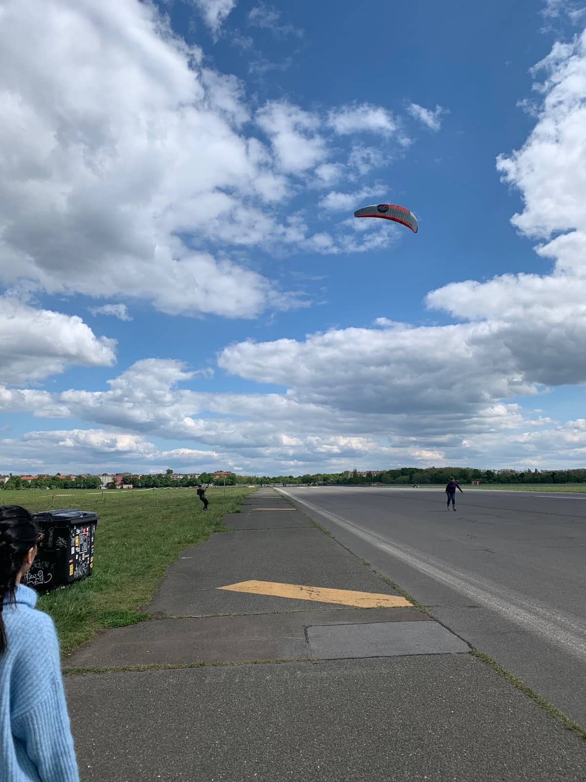 An open concrete area with someone paragliding. In the foreground to the left is a woman watching. There is a person rollerblading to the right in the distance. The skies are blue with many clouds