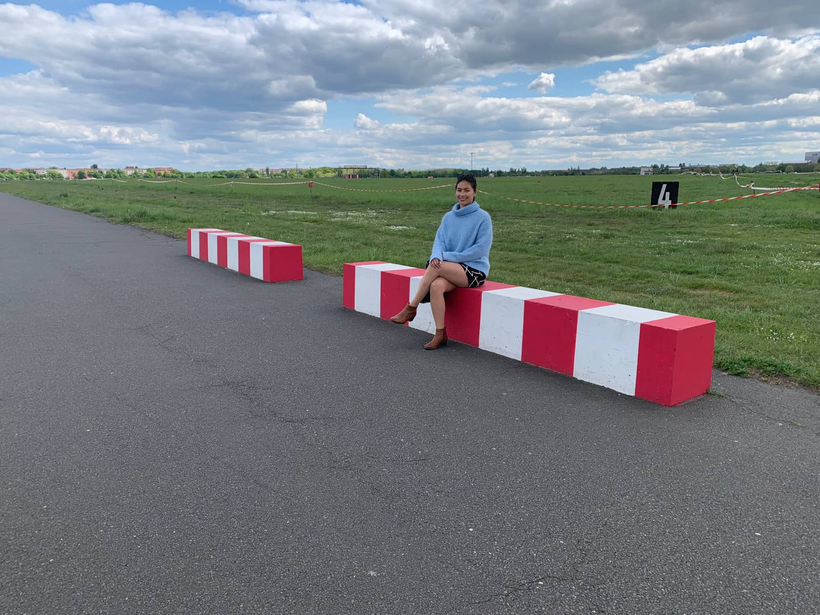 A woman on a bench-sized block of concrete painted red and white. She’s smiling with her legs crossed and is wearing a light blue sweater. In the background is a lot of grass but with areas sectioned off with tape.