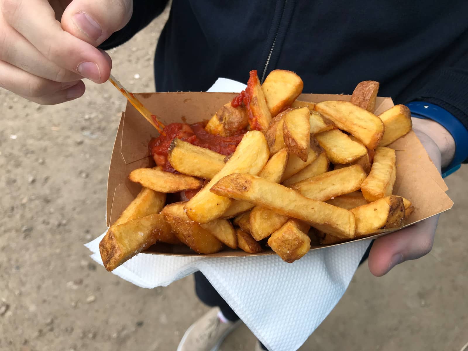 A man’s hand holding a small cardboard tray of chips, chopped sausage and sauce