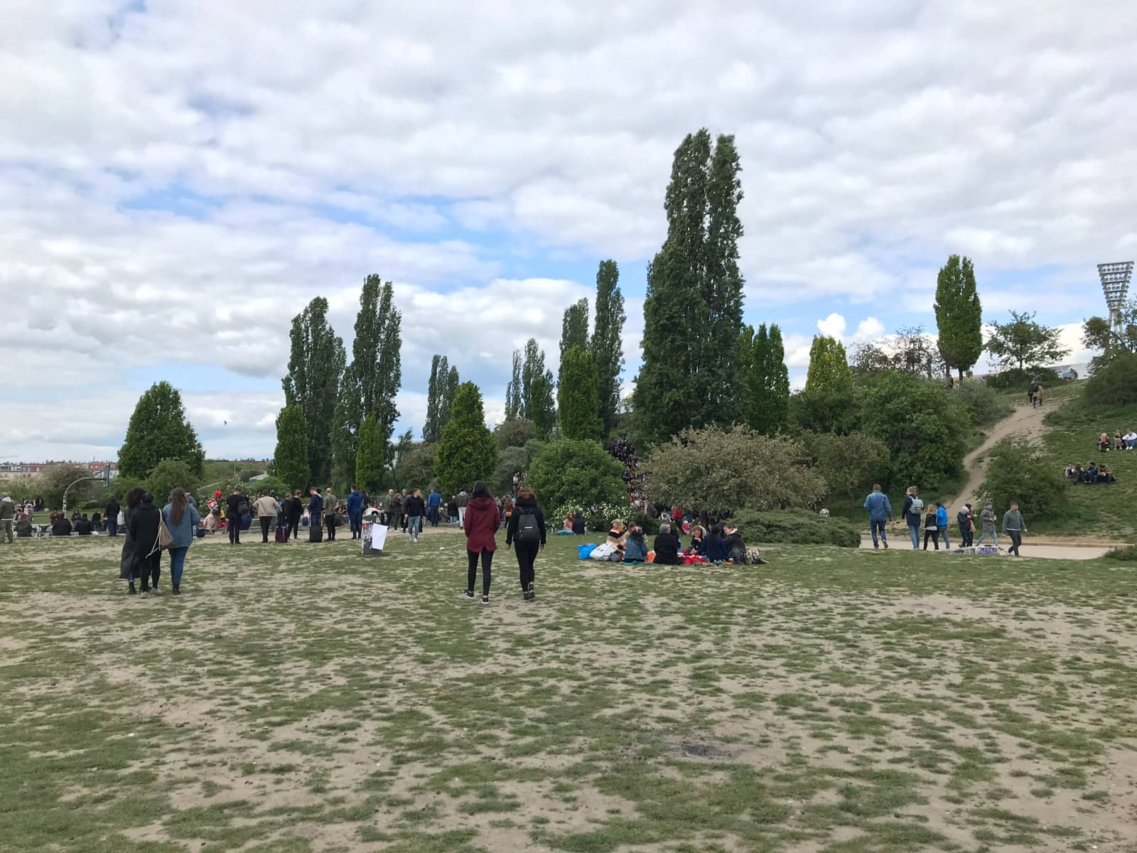 A barren area of a park where the grass is withering away, with people sitting down for a picnic and some walking