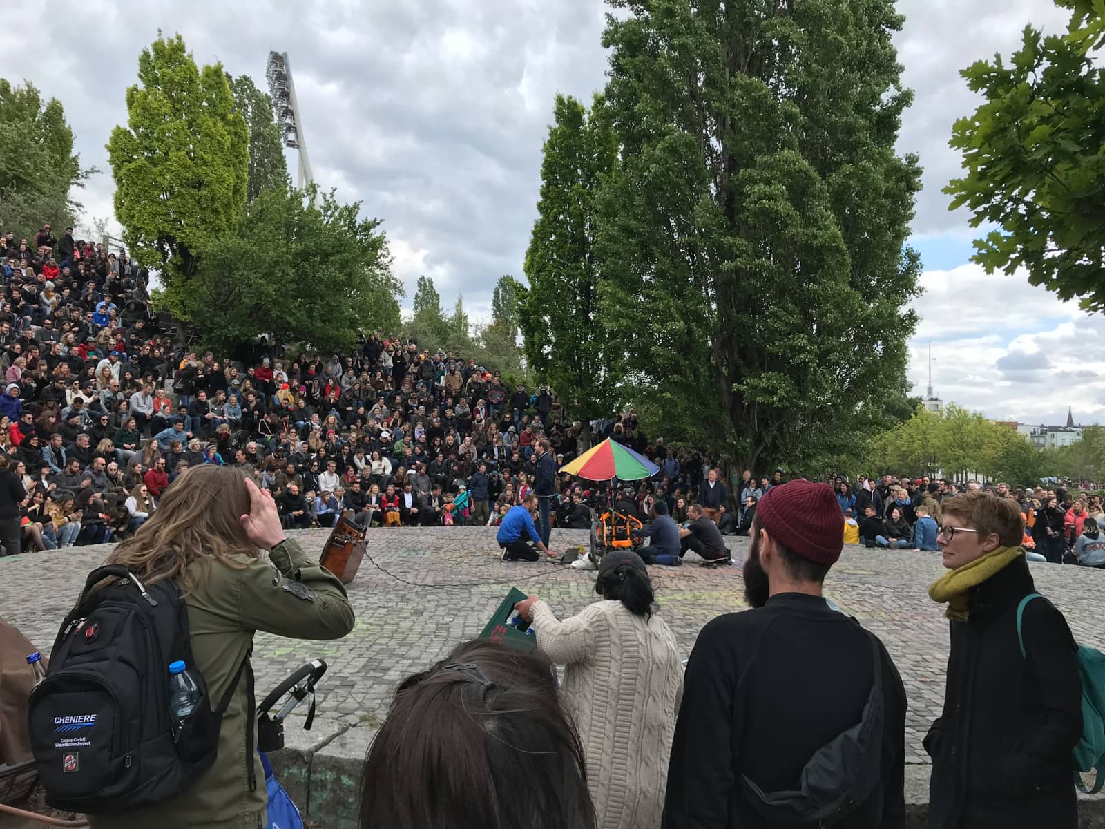 An open concrete stage with an umbrella on it and some people setting up music from a computer. To the sides and in the foreground are an audience of people watching the event