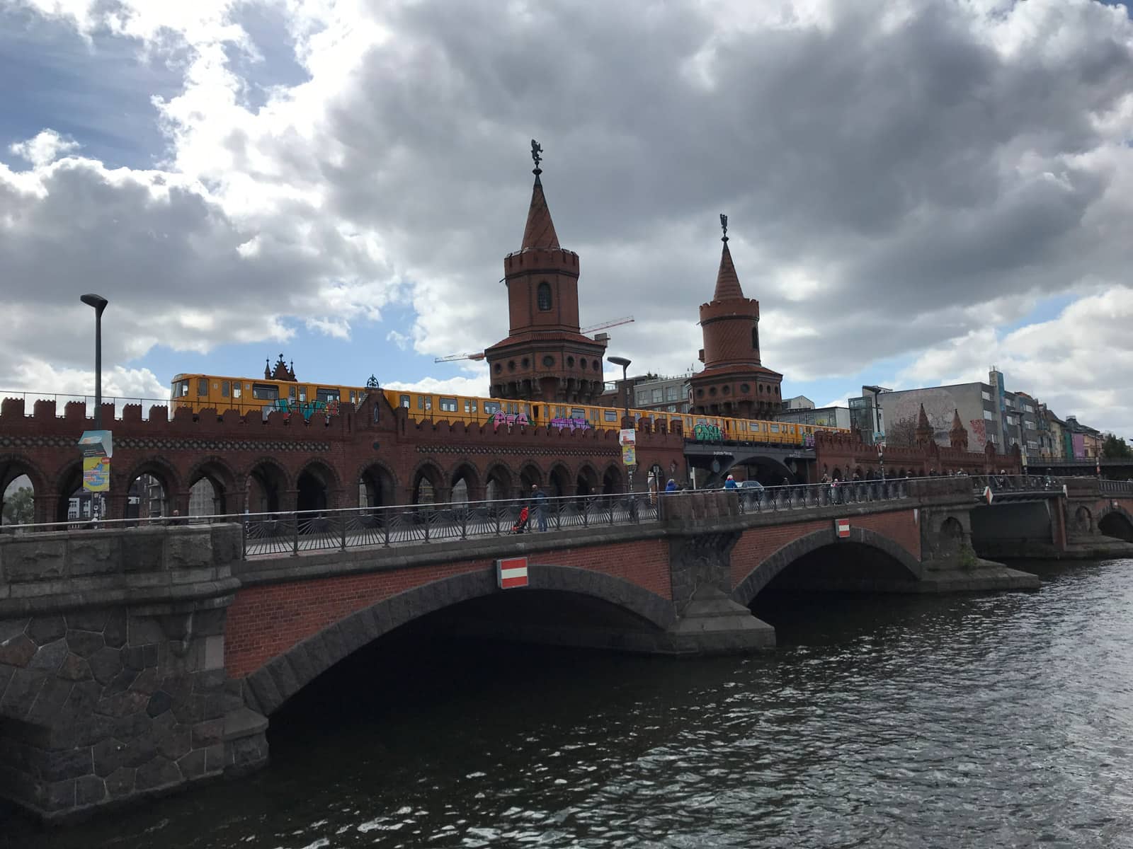 A bridge with a yellow train running across it, as seen from across the water that bridge sits on. It’s a cloudy day