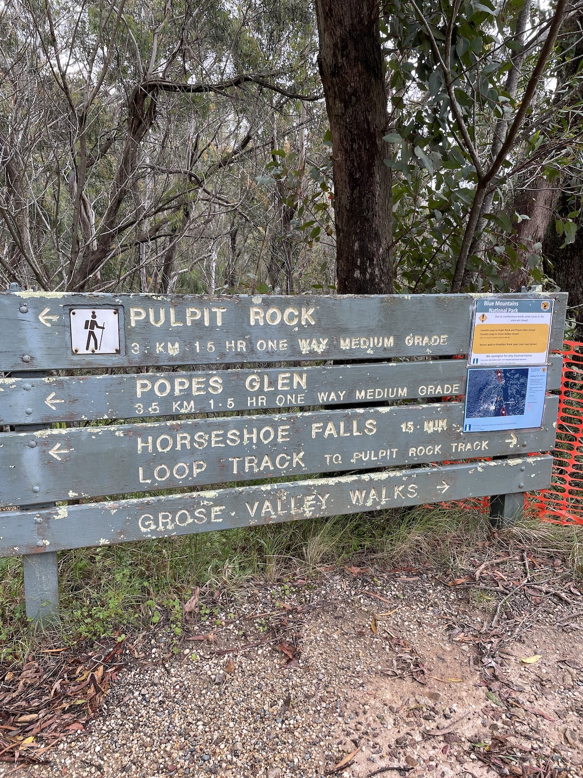 A wooden signpost showing some hiking trails, with a laminated card attached to part of the signpost, and some orange netting to the right, indicating that the area is undergoing maintenance.
