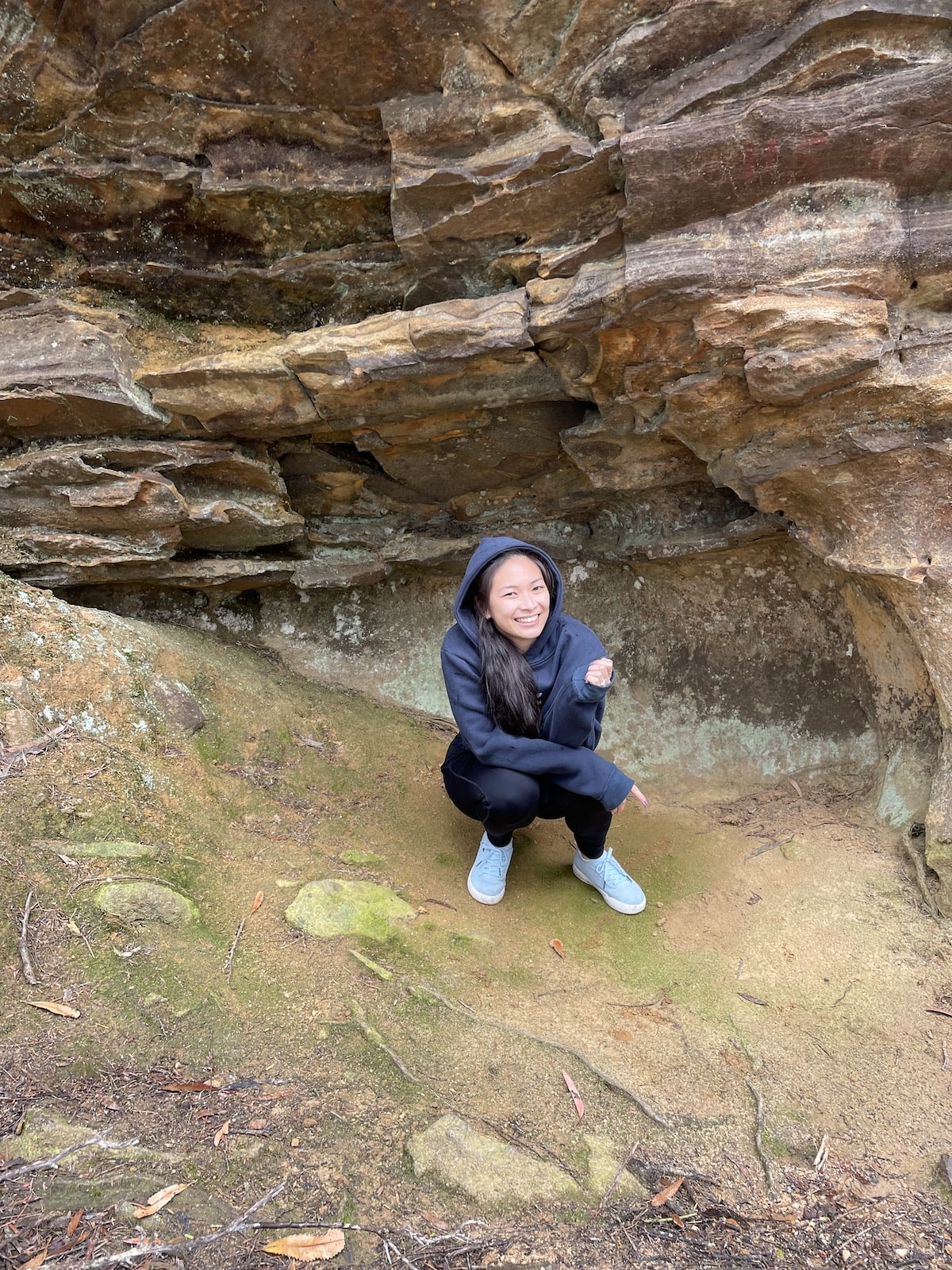 Me, Georgie, wearing a dark hoodie and pants, crouching down in a nook of a light brown coloured rock cave