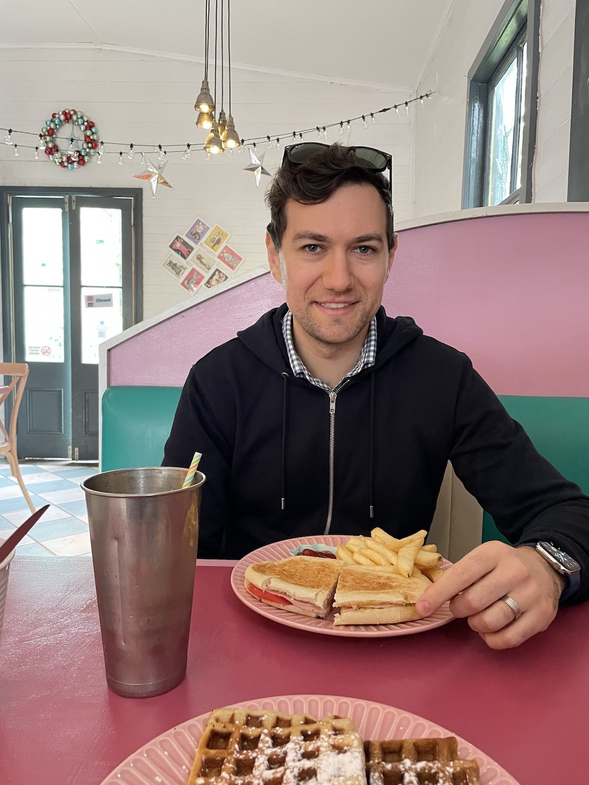 Nick, wearing a dark jacket, sitting in the pink booth of a diner with a plate of chips and a ham and cheese toastie, and a stainless steel milkshake cup. In the foreground is a plate of waffles