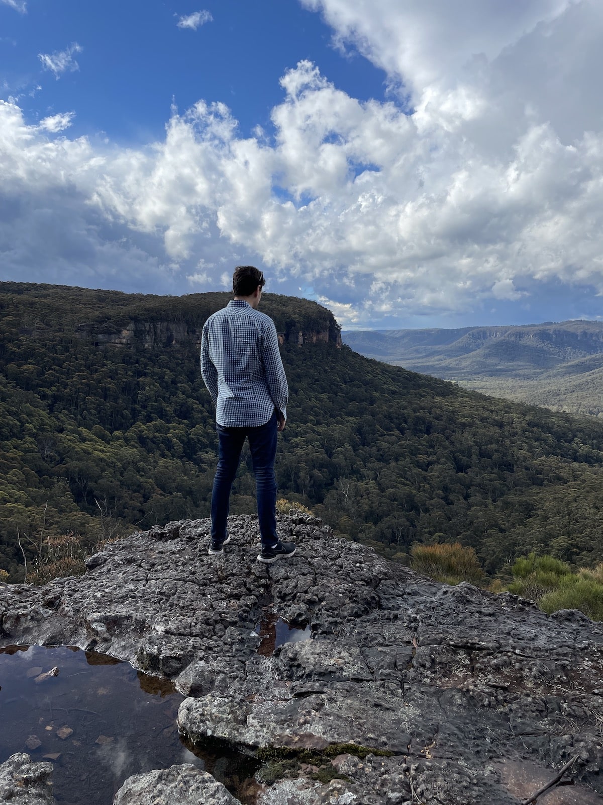 Nick, wearing a checkered shirt and dark jeans, standing at a cliff edge with green trees in the distance. The sky is bright blue with clouds. In the foreground is a small puddle of rainwater filling a hole in the rock of the cliff