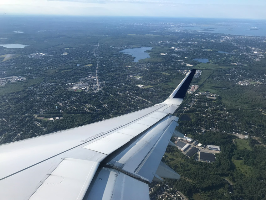 A view of a city from wing of a plane. The view is mostly green, with some major roads, and a couple of small lakes in the distance