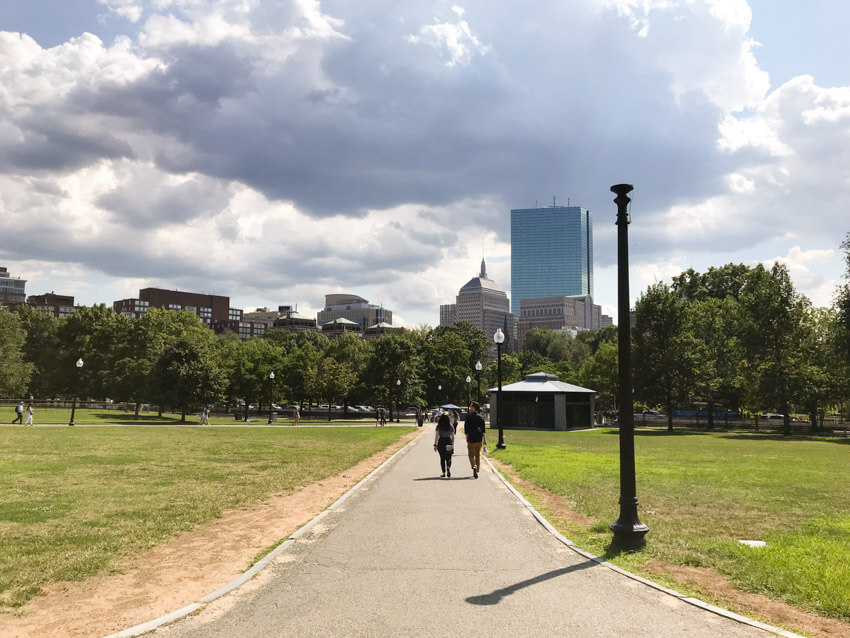 A footpath through a rather empty park, with a city in the distance. The sky is quite cloudy.