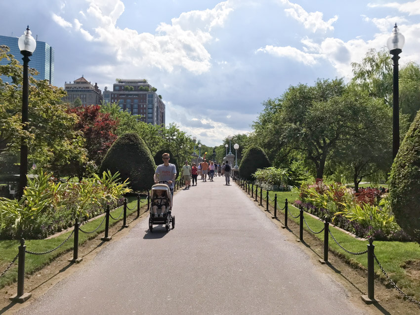 A path in a park, with some people walking on it. On the sides of the path are trees and shrubs