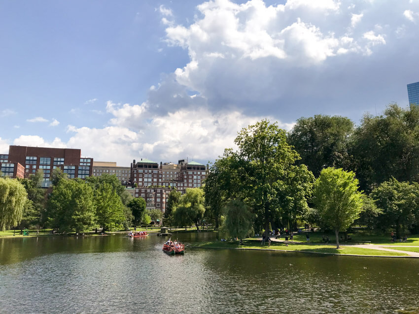 A lake in a park, filled with green trees and green grass. There is a city in the distance with brown buildings. Some boats are on the lake.