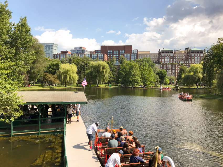 A large lake inside of a park. Down below in the foreground is a boat on the water, with wooden seats that look like benches. In the background are more of the same kind of boat. Beyond the park and its trees is a city with primarily brown buildings