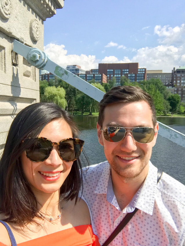 A man and woman smiling, with a lake in the background. Both of them are wearing sunglasses. The woman has short dark hair and is wearing an orange top, and the man has short brown hair and is wearing a white top with a light blue pattern.