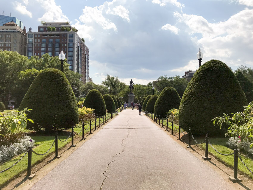 A pathway bordered on the sides by short poles with chains between the poles. There are groomed shrubs and green grass on the sides of the path. In the distance is a tall statue of a man on a horse.