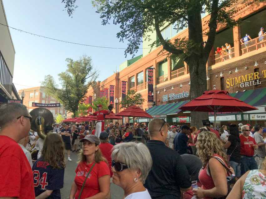 The outside area of a stadium, with many spectators lining up for food stalls and watching a man on stilts in the background. Many people are dressed in red, showing support for a baseball team.