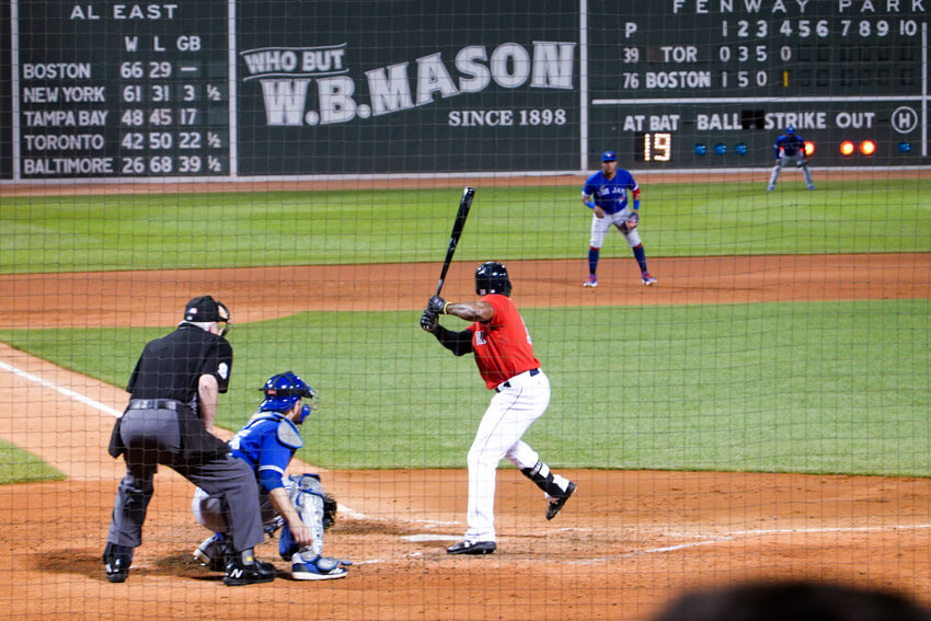 A scene from a baseball game, taken from a spectator’s point of view. A baseball player is holding a bat, ready to hit a ball. Other players and the scoreboard can be seen in the background