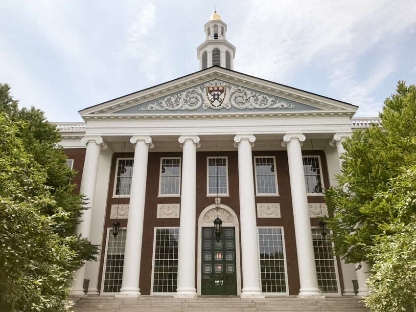 A tall building set with pillars and a triangle-shaped roof. The front is made up of tall glass windows and there is a large, tall green door in the middle