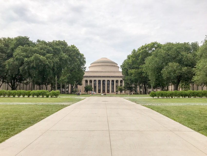 A wide path leading towards a building with many pillars and a slightly dome-shaped top. There are green trees to the sides. On the building is engraved “Massachusetts Institute of Technology”