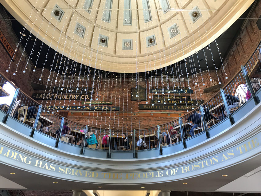 A low angle view of the inside of a building, showing upstairs seating. Some people are sitting at small tables and chatting. Hanging from the dome ceiling is a series of fairy lights.