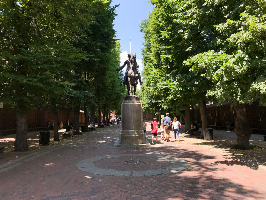 A statue of a man on a horse, in the middle of a path that is well shielded from the sun by very tall trees on the side.