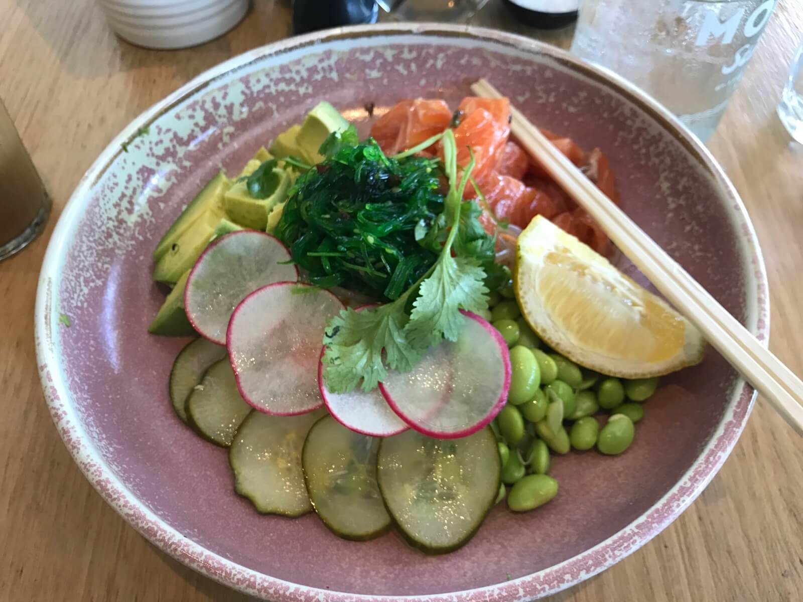A bowl with sliced pickled cucumber, edamame, seaweed, raw salmon and radish