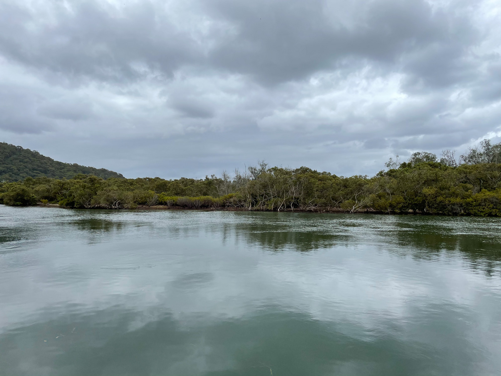 A view of a dark aqua lake with some trees on the land in the background. The sky is quite grey and cloudy, but not dark