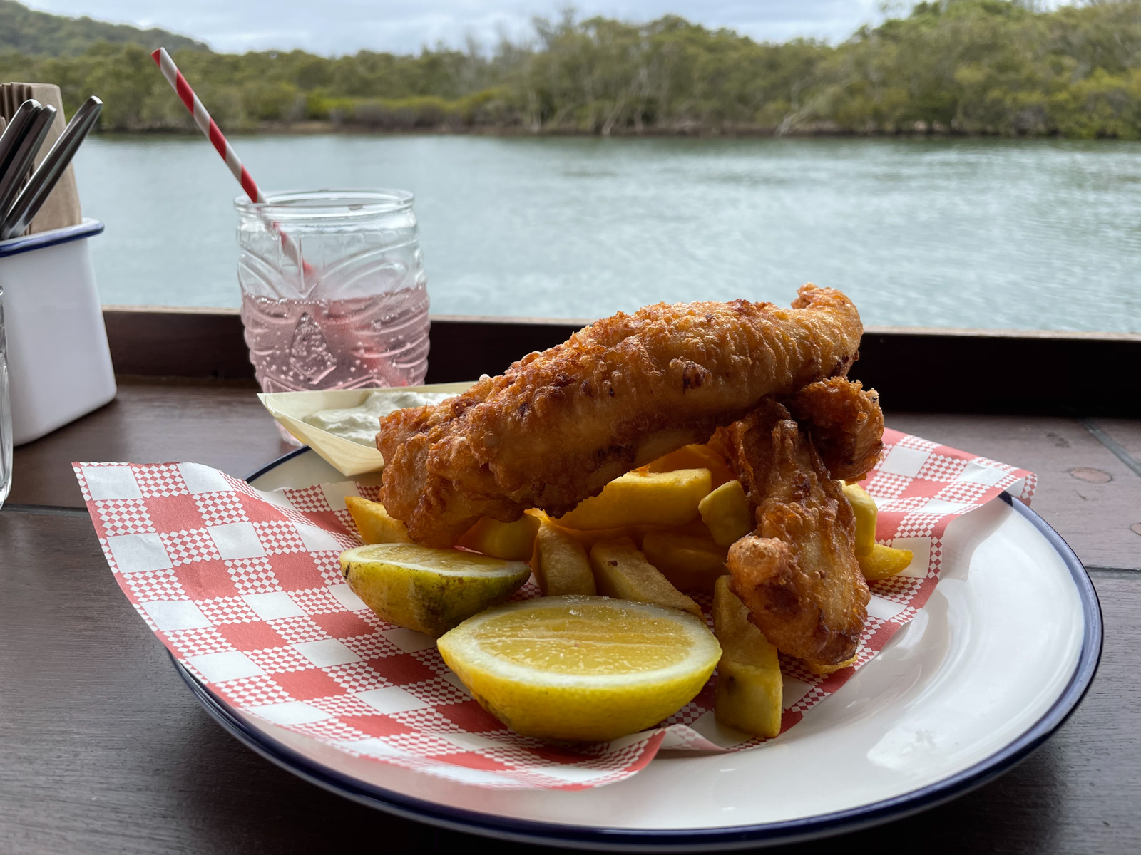 A plate of battered fish and chips on a wooden surface. Behind it is a glass with a translucent pink liquid in it. The view in the background is of a lake