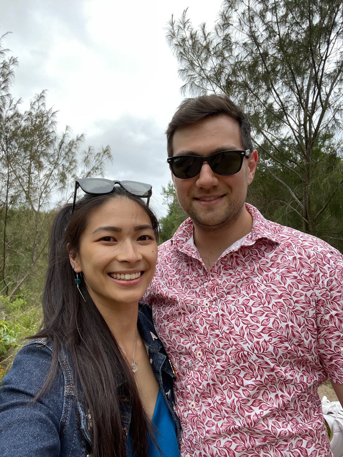 A selfie of a man and woman. The man is wearing a shirt with a red leaf pattern and the woman is wearing a blue top with a dark denim jacket. The man has dark sunglasses on and the woman has sunglasses on top of her head