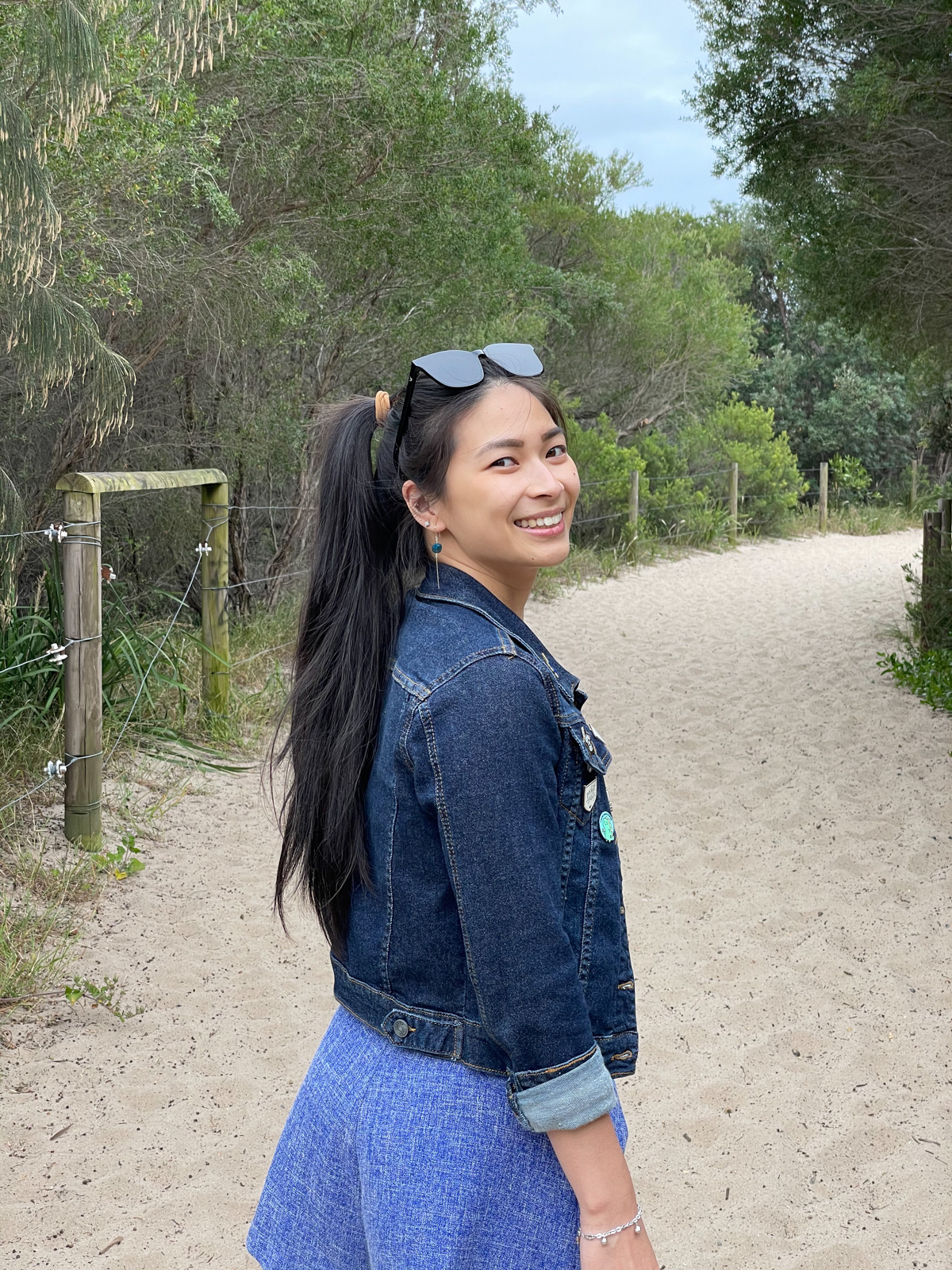 A woman with long dark hair in a ponytail, looking over her shoulder and smiling, with her right shoulder slightly tilted towards the camera. She is wearing bright blue shorts and a dark denim jacket.