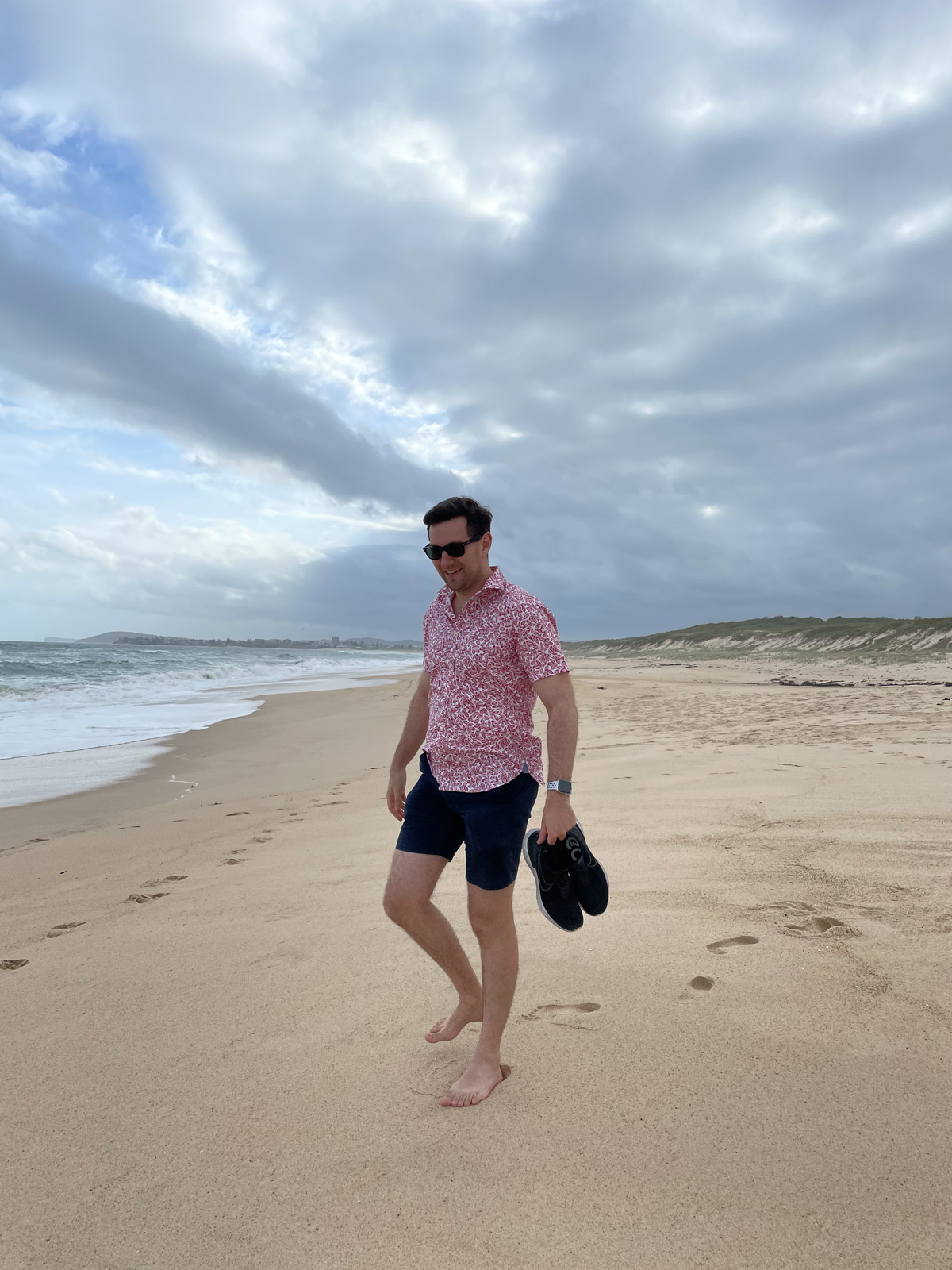 A man wearing dark sunglasses, in a red patterned shirt and dark blue shorts, standing barefoot on the sand of a beach. The sky is cloudy