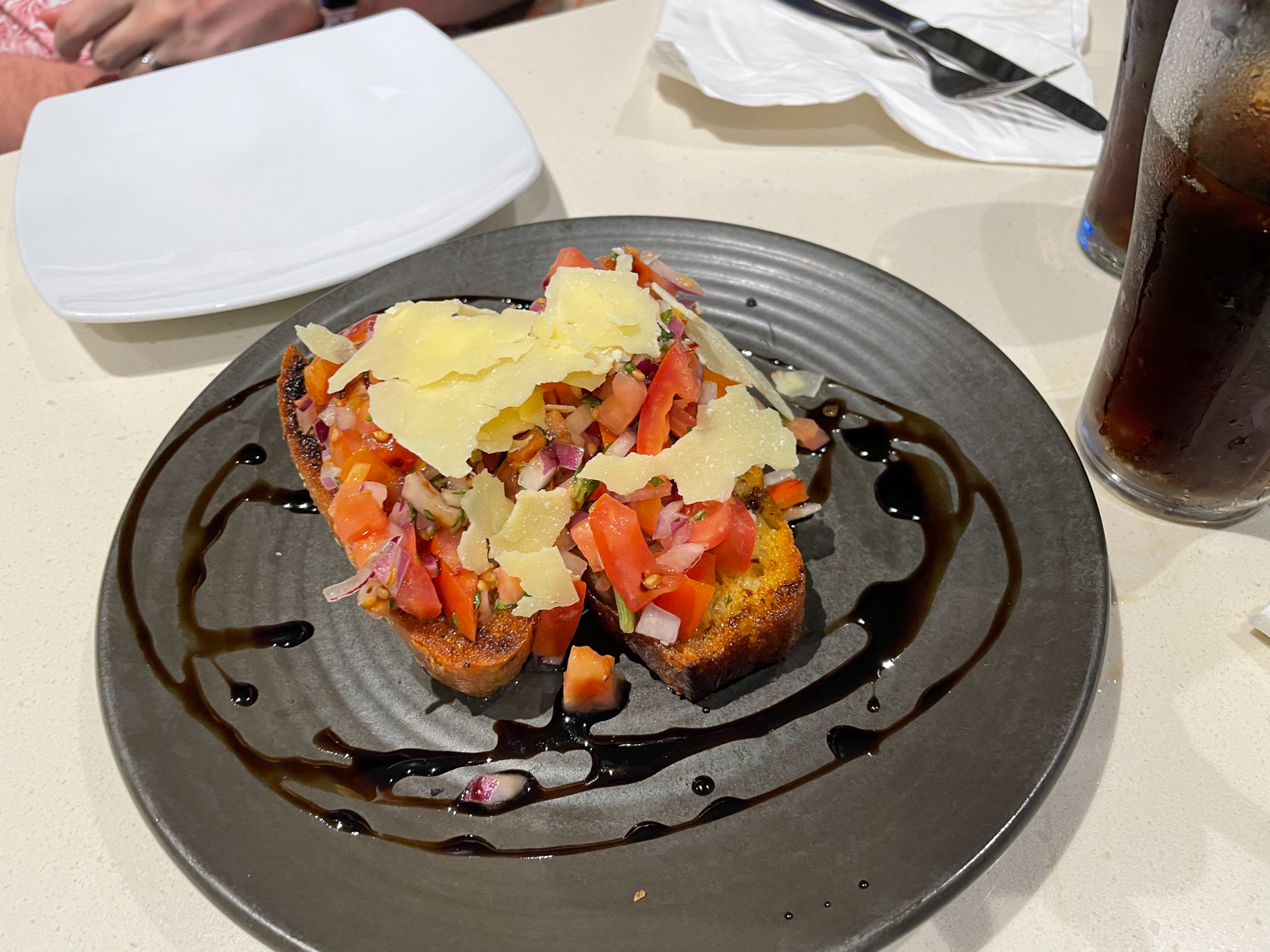 A dark coloured plate with a slice of bruschetta, topped with tomato, onion and parmesan cheese, and drizzled with balsamic vinegar glaze