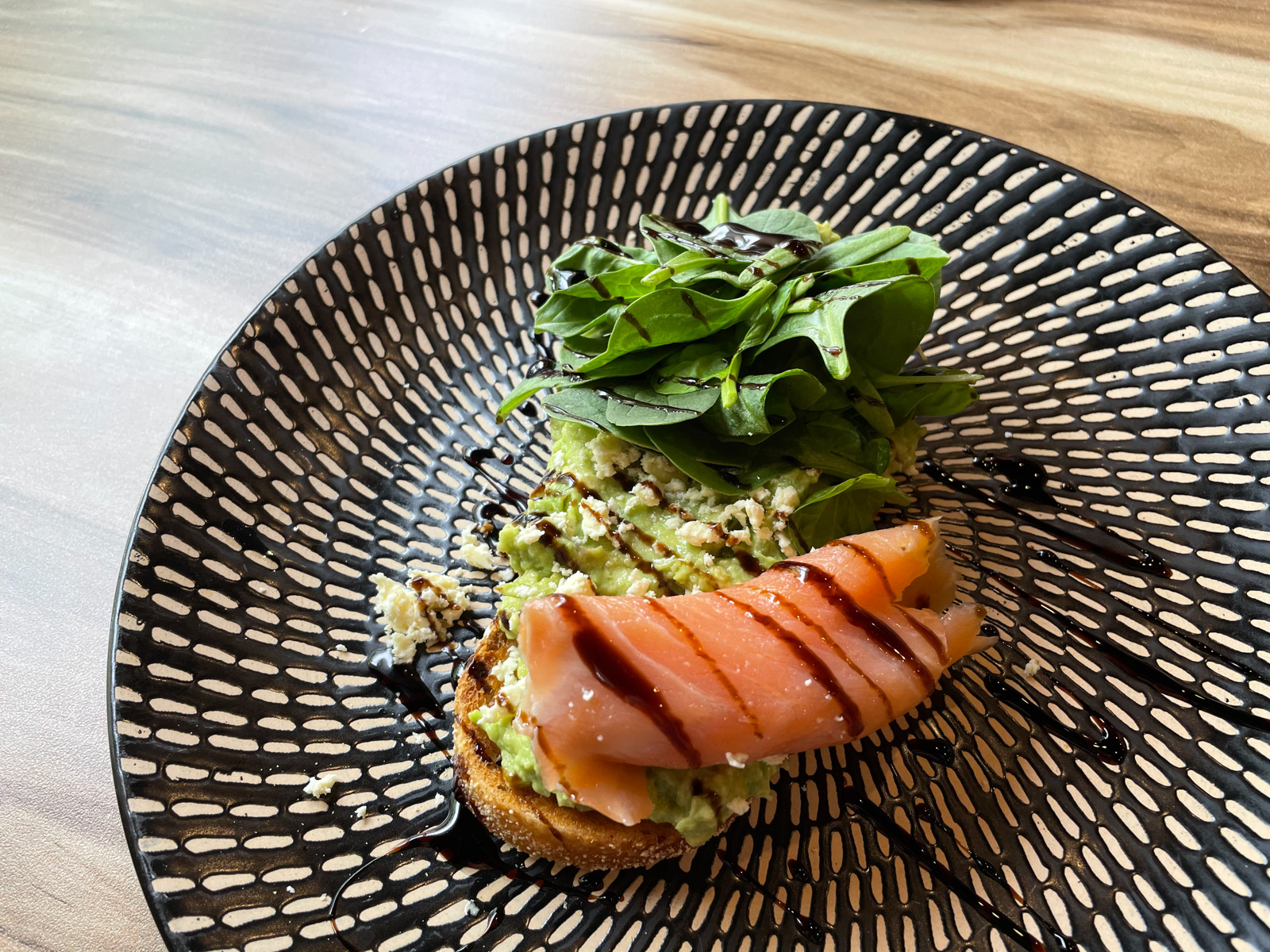 A black and white patterned plate with a slice of toast with smashed avocado spread on it, and topped with spinach, smoked salmon and balsamic glaze