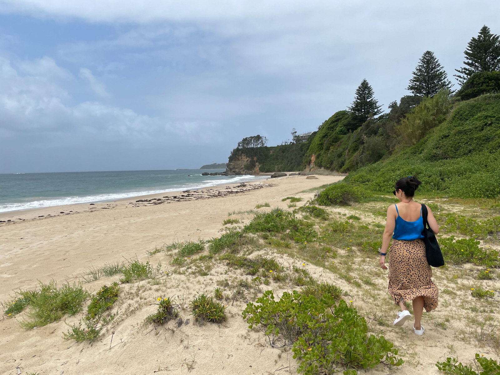 A view of a beach, with grass growing out of the sand in the foreground. A woman wearing a blue top and an animal print skirt walks towards the bare sand. The sky is a bit cloudy.