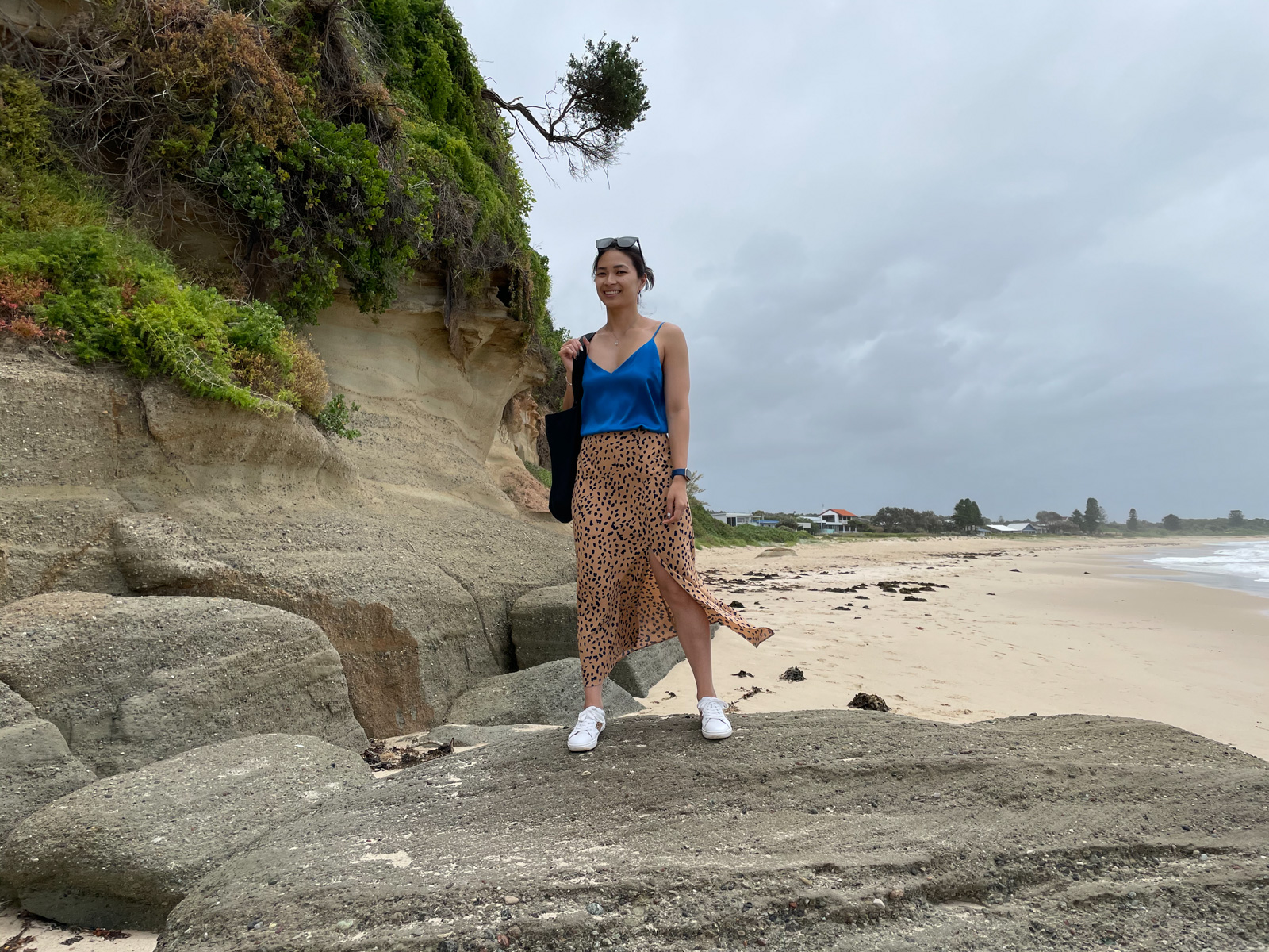 A woman wearing a blue top, animal print skirt and white shoes, standing on a giant rock on a beach. She is smiling and carrying a black tote bag, and wearing sunglasses on top of her head. The sky looks a little grey and cloudy.