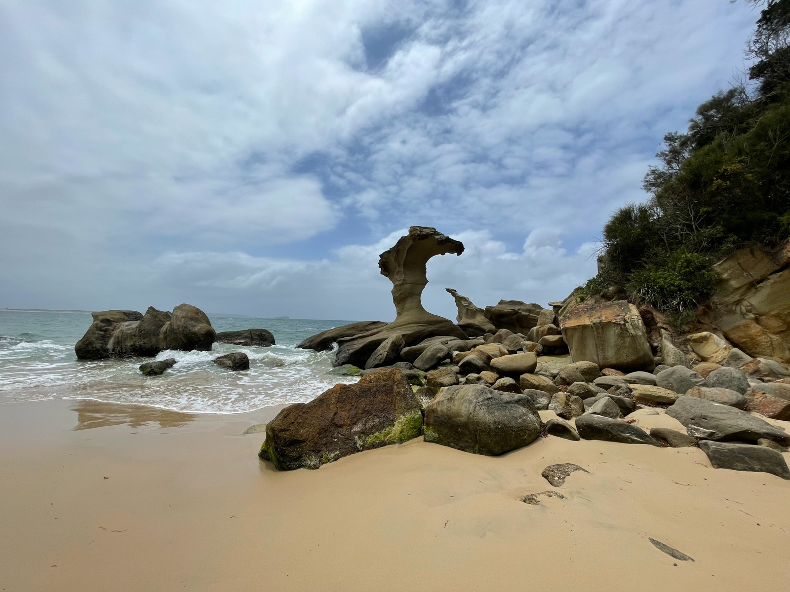 The view on a beach with some large rocks and a rock formation, one of which resembles a big wave. The sky is blue with a few clouds
