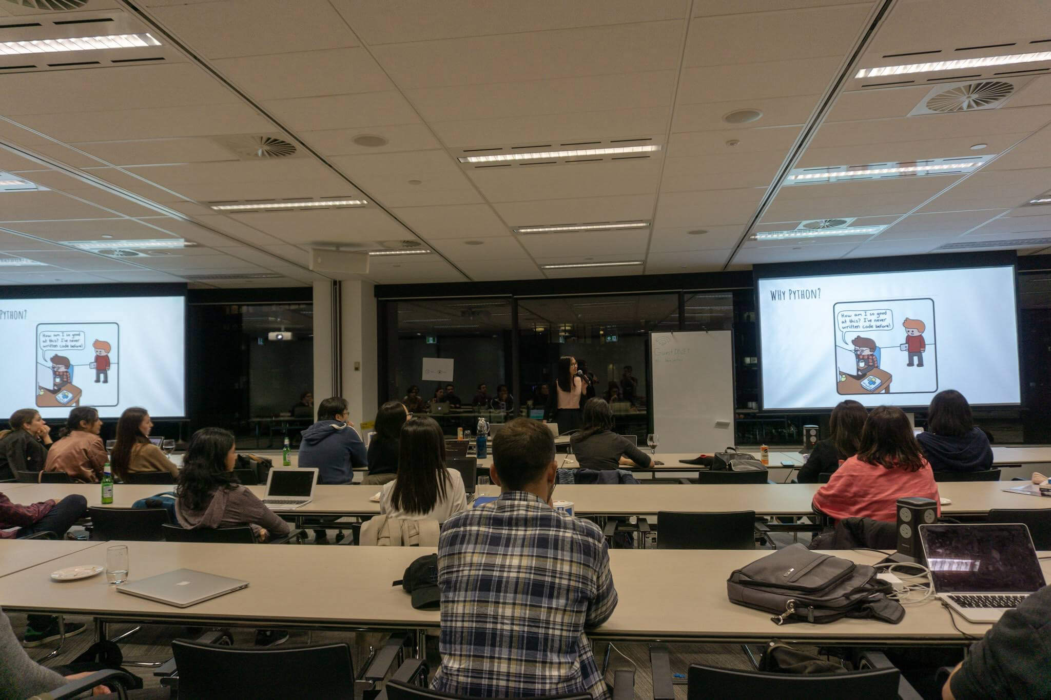 A room with rows of desks, with people seated, and a woman at the front giving a presentation alongside large screens