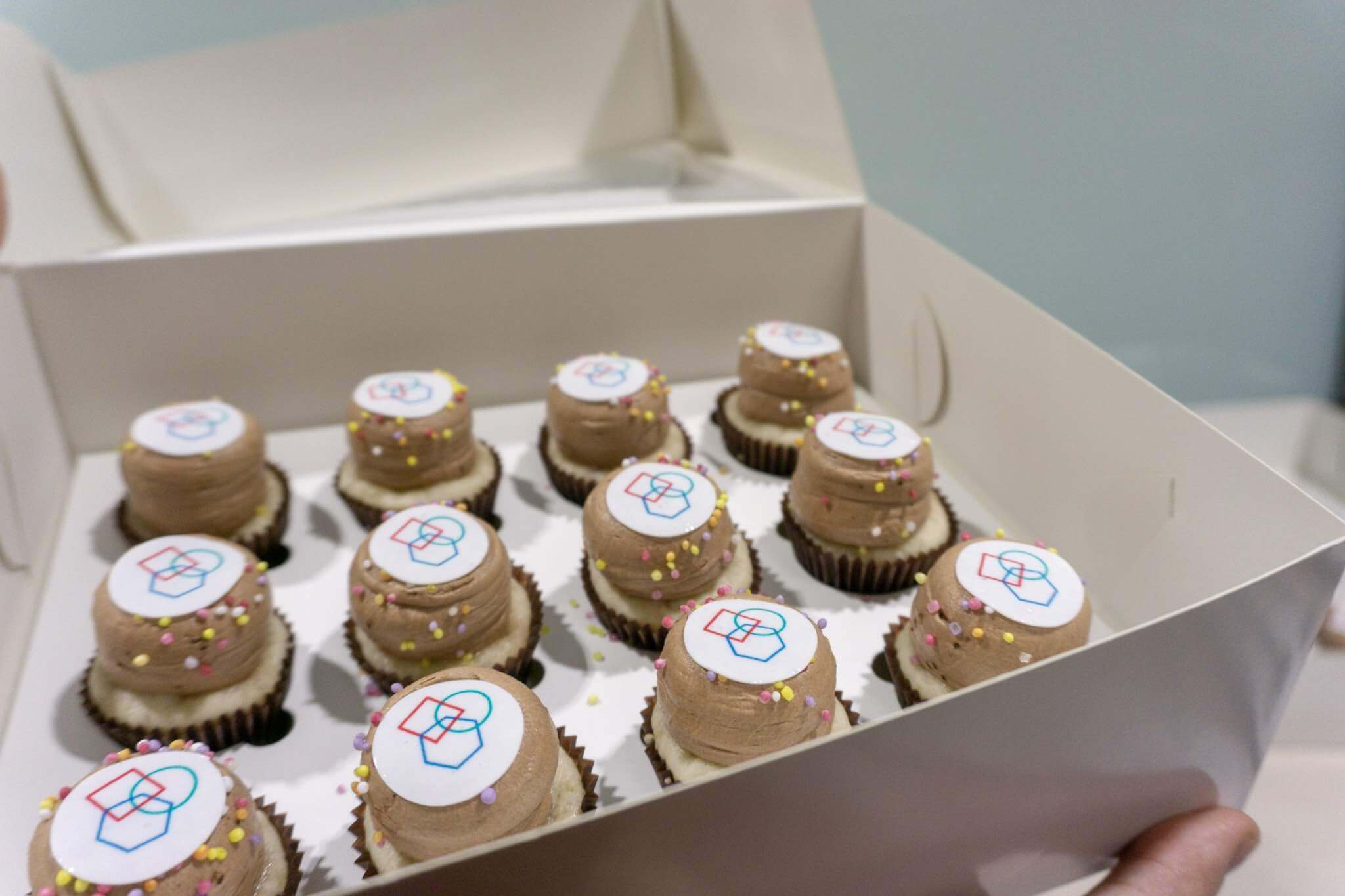 A white cardboard box with twelve miniature cupcakes. They are brown and on top have a circle with a logo on it. The logo is comprised of the overlapping outlines of shapes