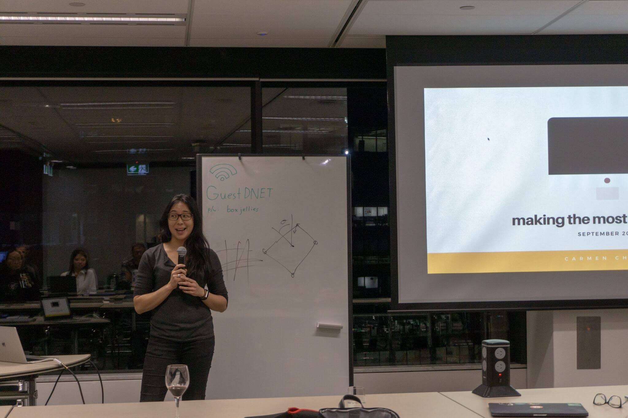 A woman wearing glasses, holding a microphone, standing next to a whiteboard and a large screen presentation