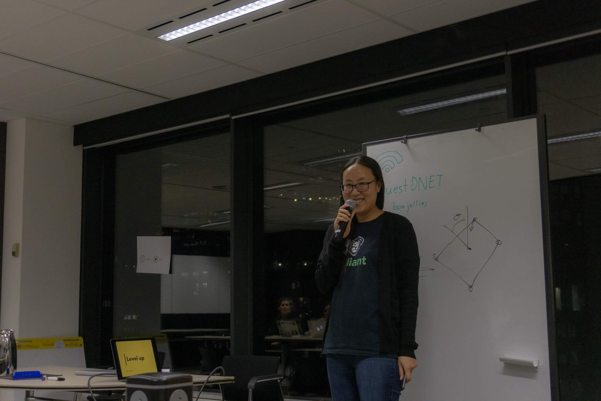 A woman wearing glasses, holding a microphone, standing next to a whiteboard