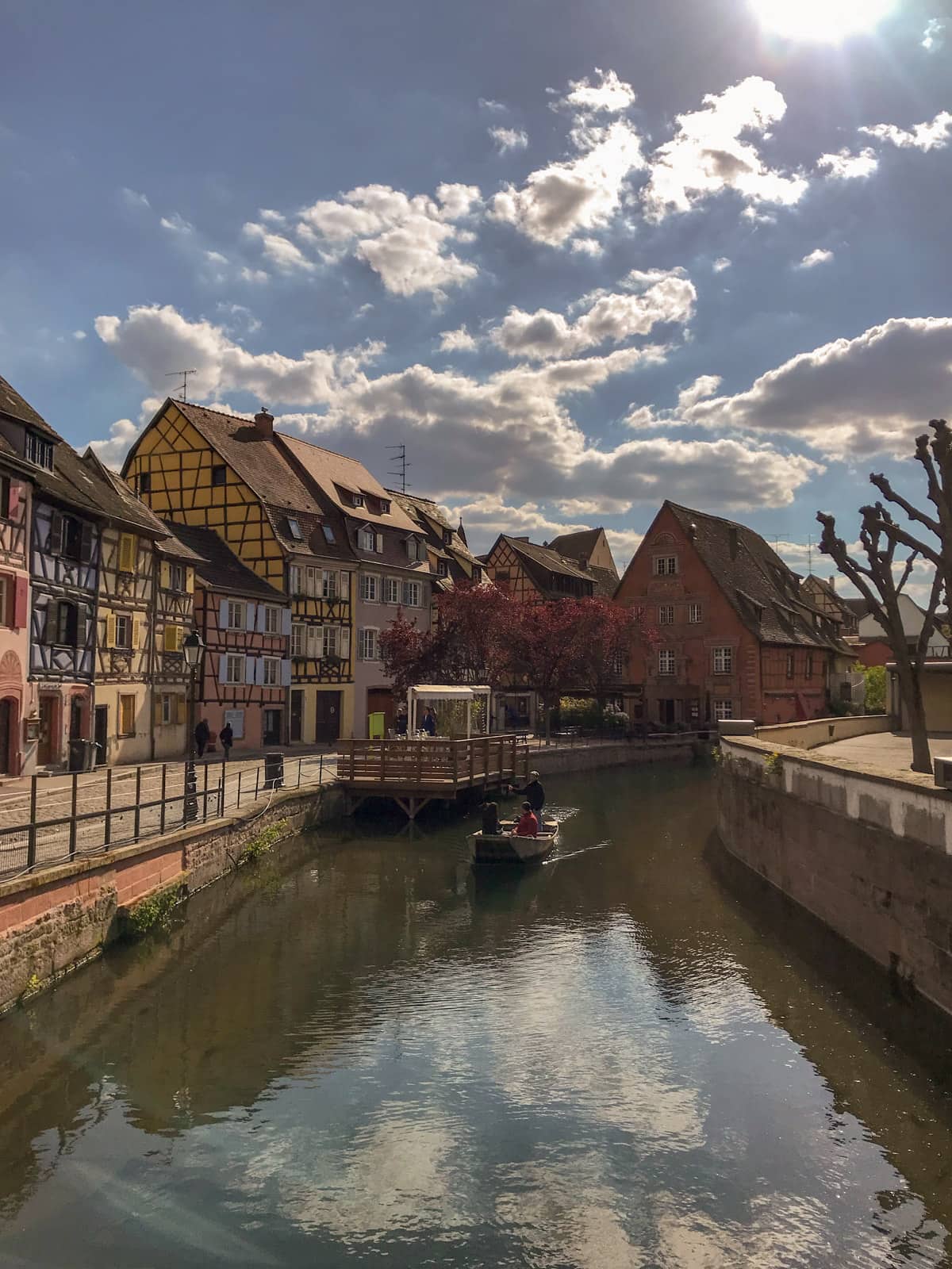A river in Colmar, France, showing some people in a small boat leaving one side of the river. The buildings on the side of the river are old and pastel-coloured but colourful
