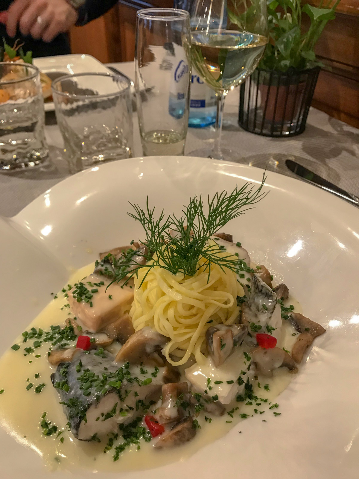 A shallow white dish with cooked fish and a ball of spaghetti, garnished with herbs and served with white sauce