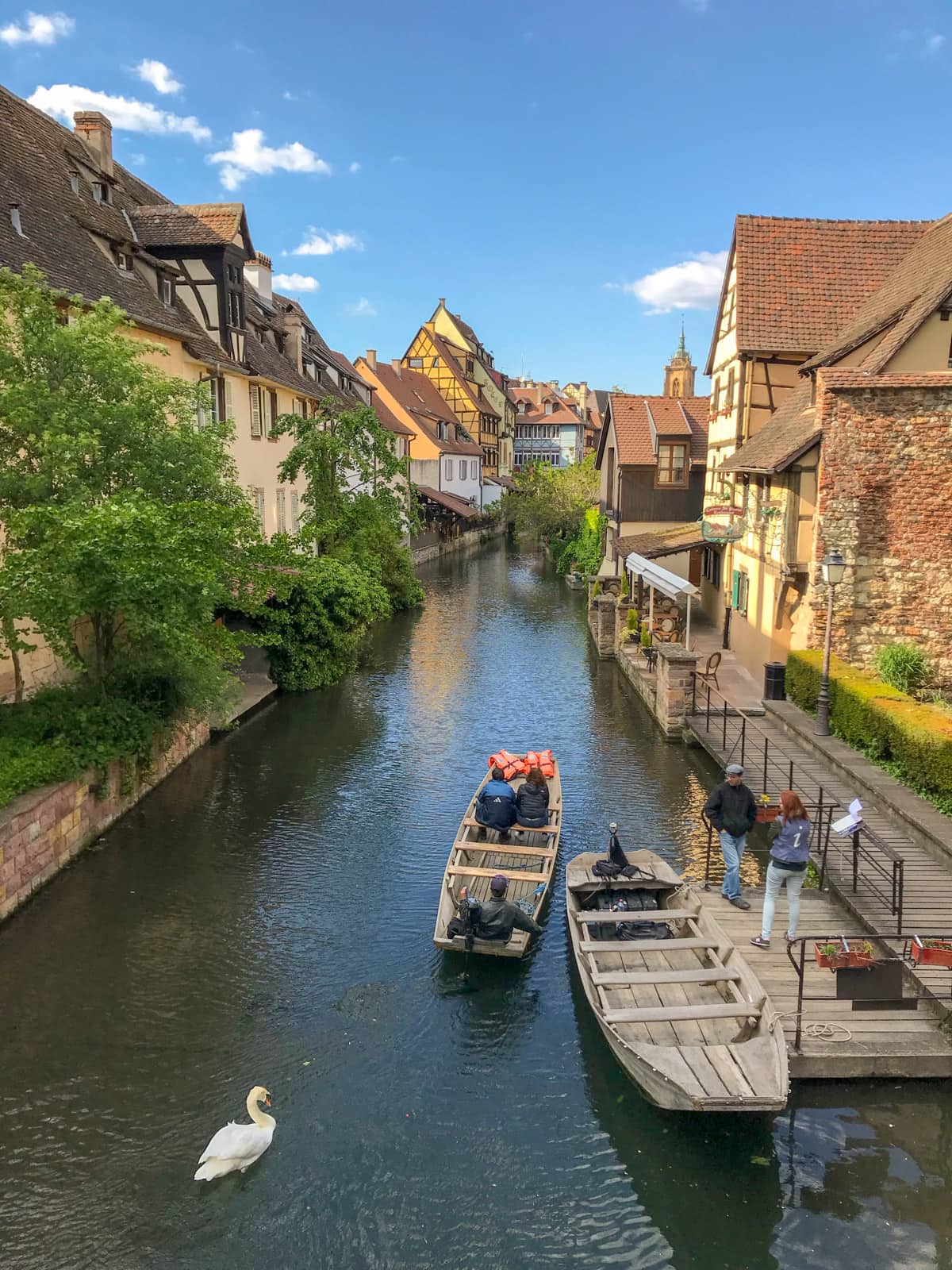 A view of a river with two wooden boats leaving a small wooden area by the side of the river. The sky is bright blue and the banks of the river have old houses built up on them